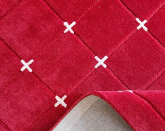 May include: A red rug with a geometric pattern of white crosses. The rug's design features a woven texture, with a portion of the rug folded back to reveal the gray backing. The rug is a vibrant home decor item.