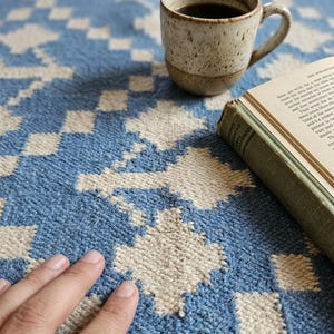 Puede incluir: Una alfombra estampada azul y blanca con una taza de caf&eacute; y un libro. La taza est&aacute; moteada con un l&iacute;quido oscuro en su interior. El libro tiene una cubierta verde y las p&aacute;ginas est&aacute;n abiertas.