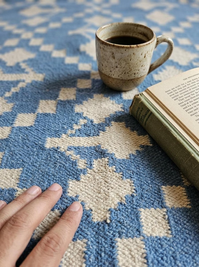 Puede incluir: Una alfombra estampada azul y blanca con una taza de caf&eacute; y un libro. La taza est&aacute; moteada con un l&iacute;quido oscuro en su interior. El libro tiene una cubierta verde y las p&aacute;ginas est&aacute;n abiertas.