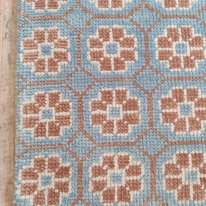 May include: A close-up of a blue rug with a repeating floral pattern in shades of brown and white. The rug has a textured surface and a fringe along the bottom edge. The floral design is geometric.