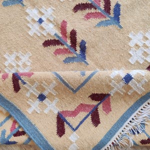 May include: A tan woven rug featuring a repeating floral pattern in white, blue, and dark red. The rug has a blue border and white fringe along the edge. The fabric shows a textured pile with a folded corner revealing the underside and fringe.