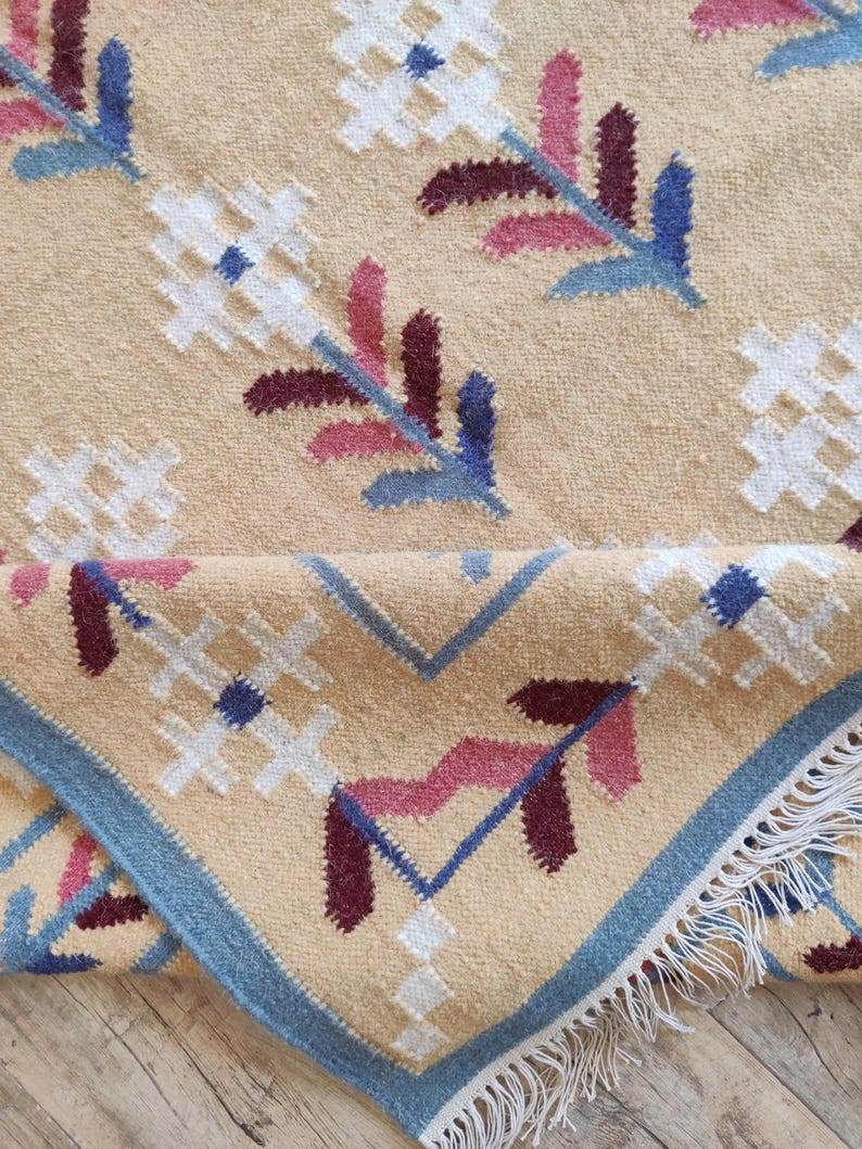 May include: A tan woven rug featuring a repeating floral pattern in white, blue, and dark red. The rug has a blue border and white fringe along the edge. The fabric shows a textured pile with a folded corner revealing the underside and fringe.