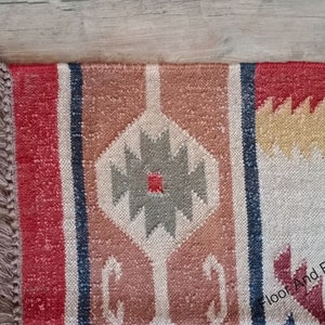 May include: A red, beige, and gray patterned rug with fringe on the edges. The rug has a geometric design with a central diamond shape.