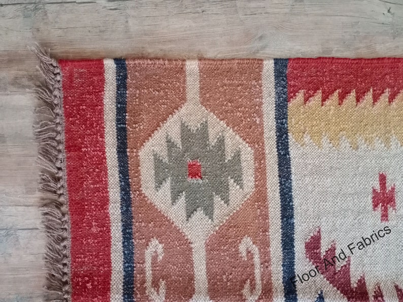 May include: A red, beige, and gray patterned rug with fringe on the edges. The rug has a geometric design with a central diamond shape.