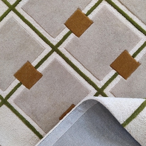 以下が含まれることがあります： 幾何学模様のカーペットのクローズアップ。カーペットは、緑と白の線で縁取られた、茶色のダイヤモンドアクセントが付いたライトグレーの正方形の繰り返しパターンが特徴です。カーペットの端が見え、灰色の裏地が明らかになっています。