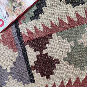 May include: A close-up of a woven rug with a geometric pattern in shades of red, brown, gray, and green. The rug has a natural, earthy feel.