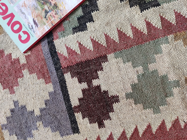 May include: A close-up of a woven rug with a geometric pattern in shades of red, brown, gray, and green. The rug has a natural, earthy feel.