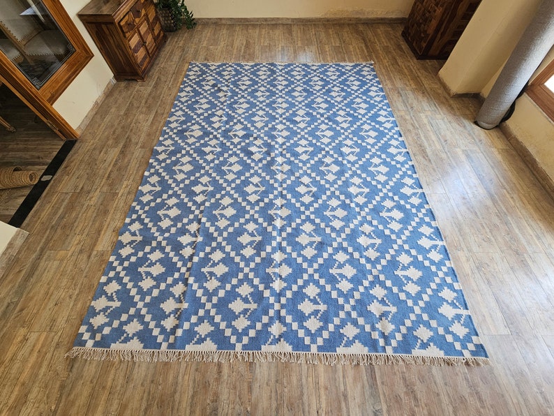 Puede incluir: Una alfombra grande y rectangular con un dise&ntilde;o geom&eacute;trico azul y blanco. La alfombra tiene flecos a lo largo del borde inferior y est&aacute; hecha de un material tejido. La alfombra est&aacute; colocada sobre un suelo de madera.