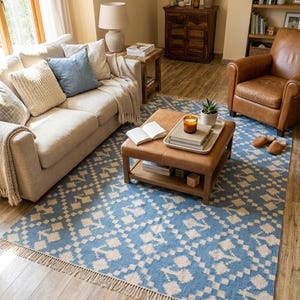 Puede incluir: Sal&oacute;n con una alfombra estampada en azul claro y crema, con flecos en el borde. La habitaci&oacute;n incluye un sof&aacute; beige con cojines, una otomana de cuero, un sill&oacute;n de cuero marr&oacute;n y muebles de madera. Una vela encendida est&aacute; sobre la otomana.
