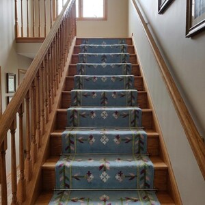 May include: A wooden staircase featuring a light blue carpet runner with a repeating floral and geometric pattern in shades of pink, green, and white. The runner is secured to each step with metallic rods, and the staircase has a wooden railing.