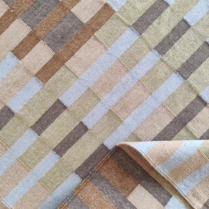 Può includere: Un tappeto rettangolare con un design patchwork. Il tappeto presenta strisce diagonali alternate in tonalità beige, marrone, grigio e azzurro. L'angolo del tappeto è ripiegato, rivelando il lato inferiore.