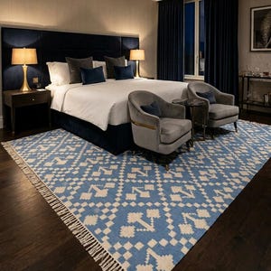 Puede incluir: Interior de dormitorio con una cama grande, cabecero azul marino y ropa de cama blanca. Dos sillones grises y una mesa peque&ntilde;a est&aacute;n colocados frente a la cama. Una alfombra estampada azul y blanca con flecos est&aacute; en el suelo. Dos l&aacute;mparas est&aacute;n en las mesitas de noche.
