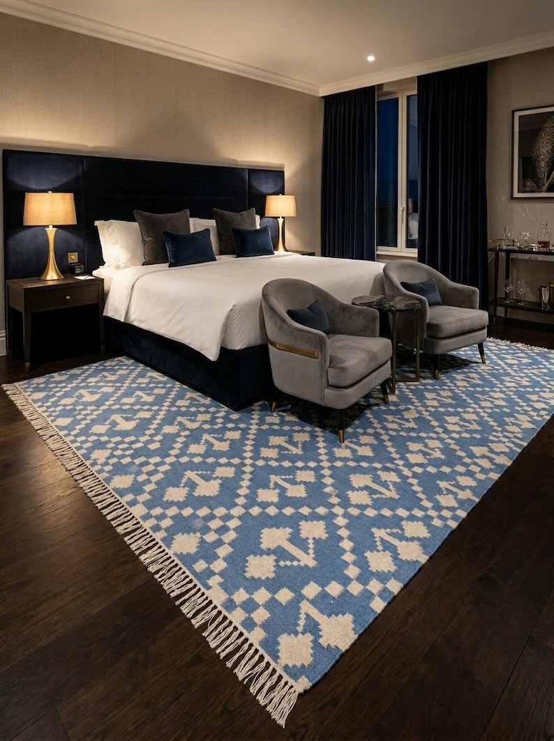 Puede incluir: Interior de dormitorio con una cama grande, cabecero azul marino y ropa de cama blanca. Dos sillones grises y una mesa peque&ntilde;a est&aacute;n colocados frente a la cama. Una alfombra estampada azul y blanca con flecos est&aacute; en el suelo. Dos l&aacute;mparas est&aacute;n en las mesitas de noche.