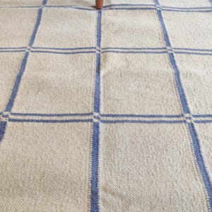 May include: A woven area rug featuring a cream-colored base with a grid pattern formed by thick blue lines. The textile has a textured surface and is partially covered by the wooden legs of a piece of furniture at the top of the frame.