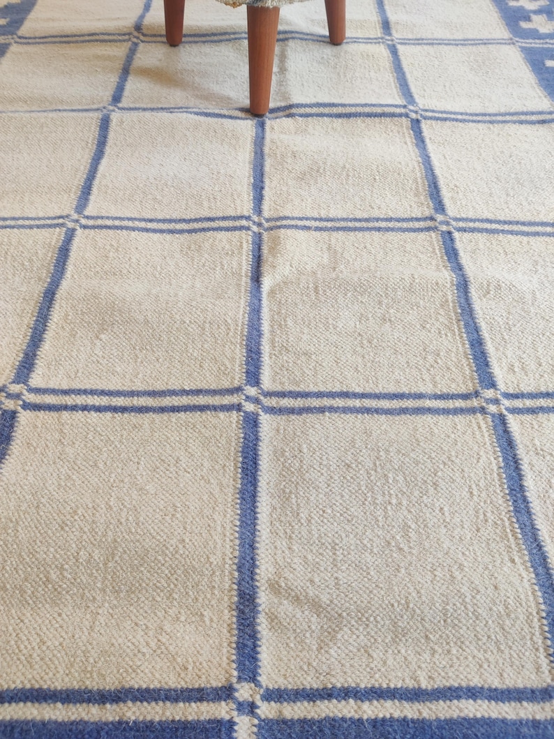 May include: A woven area rug featuring a cream-colored base with a grid pattern formed by thick blue lines. The textile has a textured surface and is partially covered by the wooden legs of a piece of furniture at the top of the frame.
