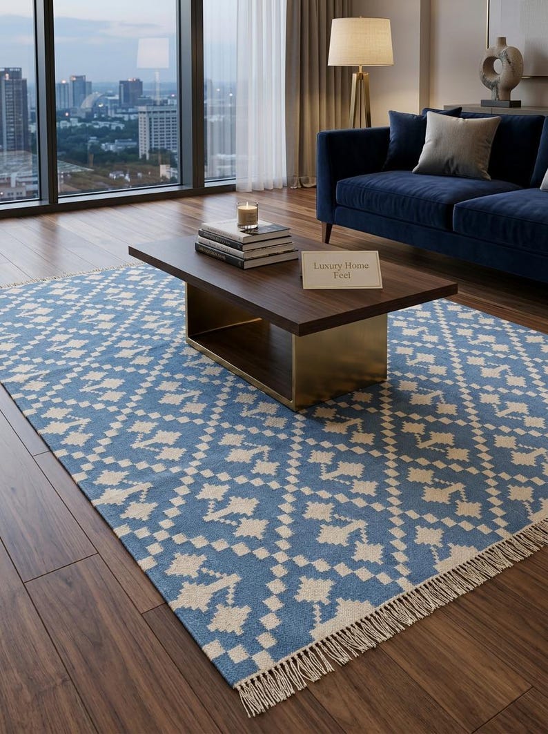 Puede incluir: Una alfombra azul y crema con un patr&oacute;n geom&eacute;trico y flecos, colocada sobre un suelo de madera. Una mesa de centro de madera oscura con detalles dorados y un letrero que dice "Luxury Home Feel" se encuentra sobre la alfombra. Un sof&aacute; azul est&aacute; en el fondo.