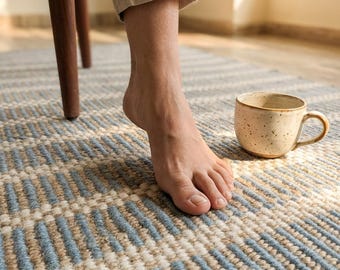 Puede incluir: Una alfombra estampada en beige y azul con una taza de cerámica. La alfombra presenta un diseño geométrico con rayas beige y azules alternas. Un pie está cerca de la taza y se ve una pata de silla de madera.