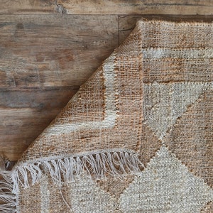 Pu&ograve; includere: Un tappeto tessuto beige e bianco con un motivo geometrico e frange. Il tappeto &egrave; piegato a met&agrave; e le frange sono visibili sul bordo inferiore.