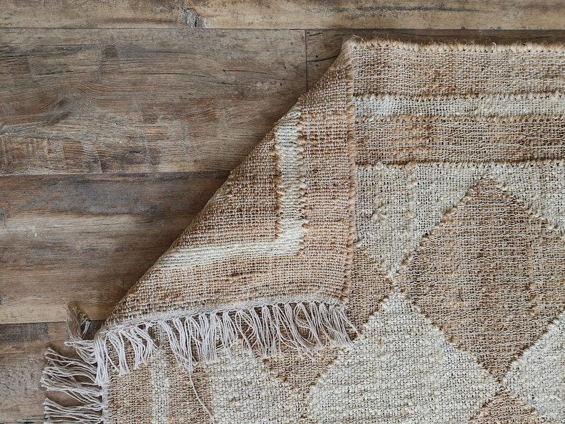 Pu&ograve; includere: Un tappeto tessuto beige e bianco con un motivo geometrico e frange. Il tappeto &egrave; piegato a met&agrave; e le frange sono visibili sul bordo inferiore.