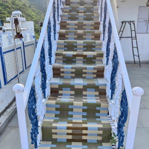 Puede incluir: Escalera pintada de blanco con barandillas de metal azul y una alfombra estampada en los peldaños. La escalera está al aire libre y tiene una vista de una colina verde en el fondo.