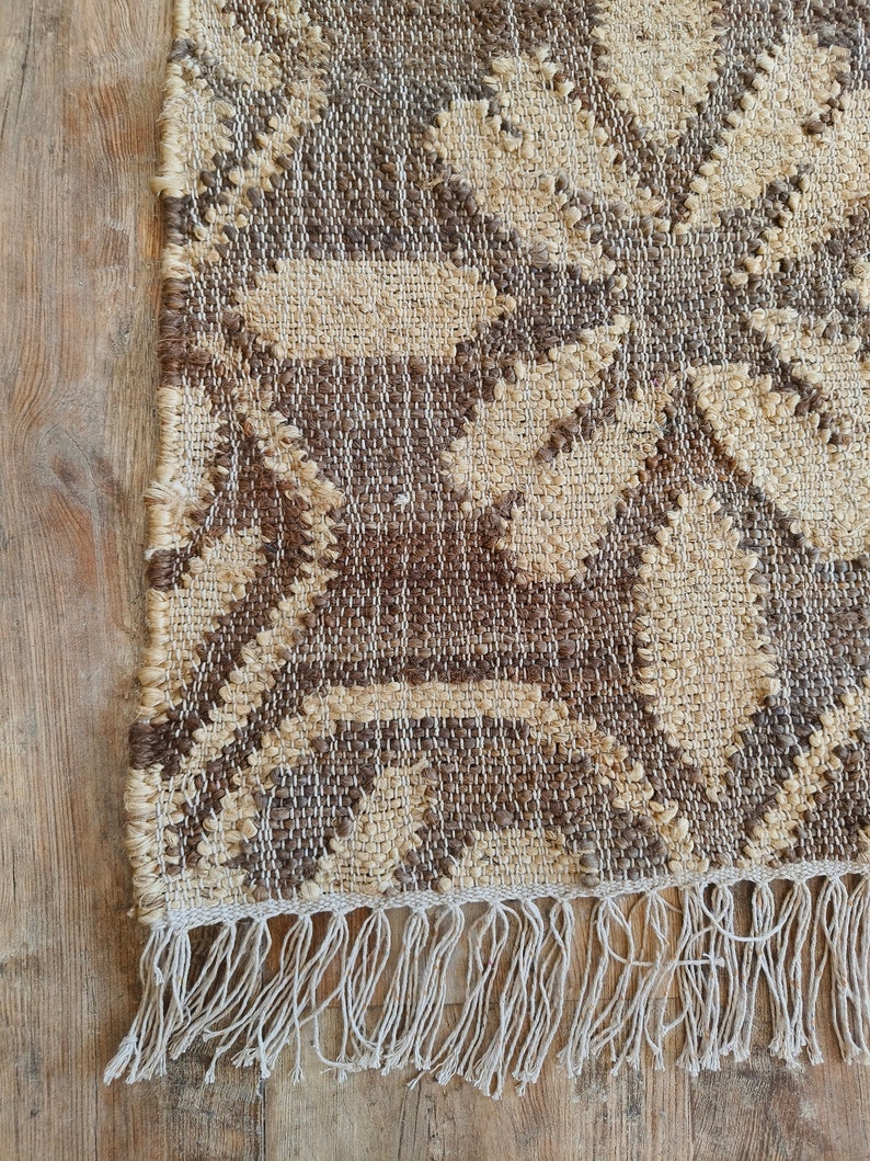 May include: A brown and beige woven rug with a geometric pattern and fringe on the bottom edge.