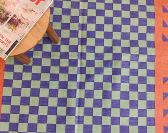 Może przedstawiać: Dywan w kratkę w kolorze zielonym i fioletowym z czerwono-pomarańczową obwódką. Dywan ma geometryczny wzór trójkątów wzdłuż obwódki.
