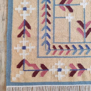 May include: A woven rug with a beige base featuring a geometric border pattern in shades of blue, white, and maroon. The design includes repeating cross motifs and arrow-like shapes, finished with a white fringe along the bottom edge.