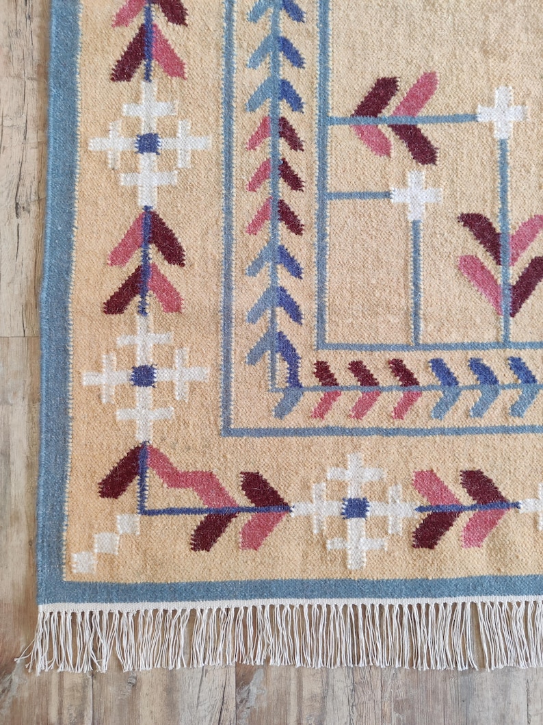 May include: A woven rug with a beige base featuring a geometric border pattern in shades of blue, white, and maroon. The design includes repeating cross motifs and arrow-like shapes, finished with a white fringe along the bottom edge.