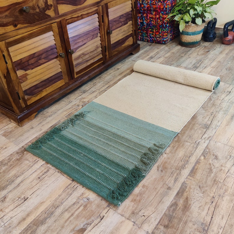 May include: A green and beige woven rug with fringed edges. The rug is rolled up and has a light beige color on the top half and a darker green color on the bottom half.