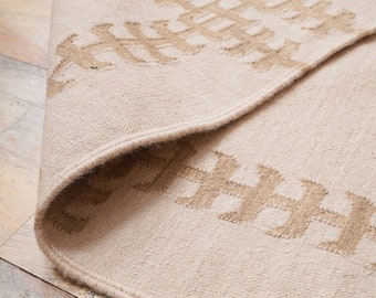 Puede incluir: Una alfombra beige con un patrón tejido de formas geométricas marrones. La alfombra tiene un borde de flecos.