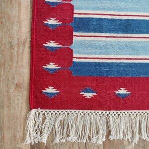 May include: A red, blue, and white striped rug with a fringe border. The rug has a geometric pattern in the border.
