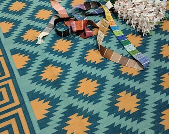 Puede incluir: Una alfombra estampada en color turquesa y naranja con un diseño geométrico. La alfombra presenta un patrón repetitivo de zigzags y formas de estrella. Una cinta métrica colorida y un ramo de flores blancas están sobre la alfombra.