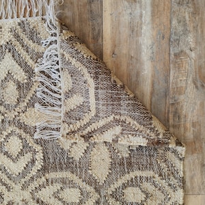 May include: A brown and beige patterned rug with fringe on the edges. The rug is folded over on a wooden floor.