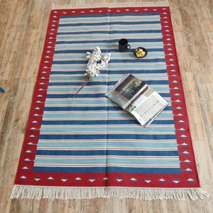 May include: A blue and white striped rug with a red border and white fringe. The rug has a geometric pattern in the border. The rug is on a wooden floor.