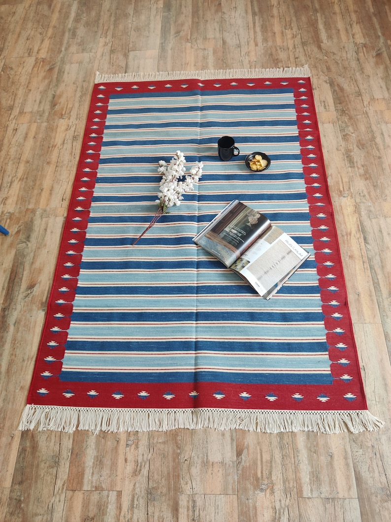 May include: A blue and white striped rug with a red border and white fringe. The rug has a geometric pattern in the border. The rug is on a wooden floor.