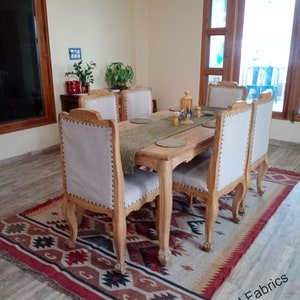 May include: A wooden dining table with six chairs, set with placemats and silverware. The table is surrounded by a patterned rug with red, brown, and black accents. The room has a light beige wall and a large window with wooden blinds.