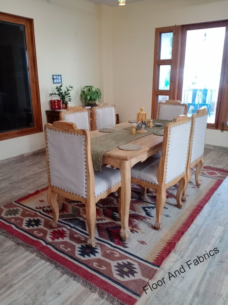 May include: A wooden dining table with six chairs, set with placemats and silverware. The table is surrounded by a patterned rug with red, brown, and black accents. The room has a light beige wall and a large window with wooden blinds.