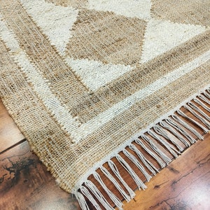 Pu&ograve; includere: Un tappeto in fibra naturale con un motivo geometrico beige e bianco. Il tappeto ha un bordo a frangia.