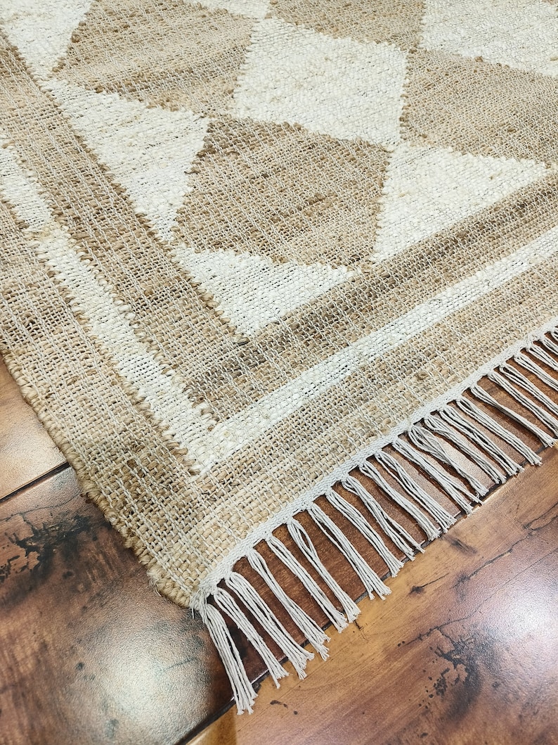 Pu&ograve; includere: Un tappeto in fibra naturale con un motivo geometrico beige e bianco. Il tappeto ha un bordo a frangia.