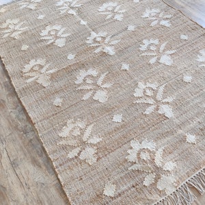 Peut inclure: Un tapis en fibres naturelles avec un motif tissé de fleurs blanches sur un fond beige. Le tapis a des franges sur tous les côtés.