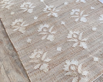 May include: A natural fiber rug with a woven design of white flowers on a beige background. The rug has fringe on all sides.