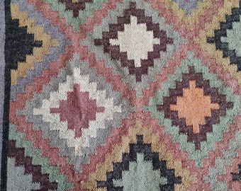 May include: A colorful rug with a geometric pattern in shades of brown, red, beige, and green. The rug has a woven texture and a diamond-shaped design.
