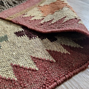 May include: A woven rug with a geometric pattern in shades of brown, beige, and red. The rug has a fringed edge and is made of natural fibers.