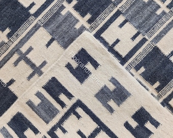 May include: A close-up of a woven rug with a geometric pattern. The rug features a repeating design of dark blue and light blue shapes against a cream-colored background. The rug is folded over, revealing the underside.