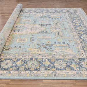 以下が含まれることがあります： ライトブルーの中央フィールドとダークブルーのボーダーを備えた、丸められた長方形のラグ。ラグは、クリーム、イエロー、グリーンの色合いで複雑な幾何学模様が特徴です。ラグは木の床にあります。