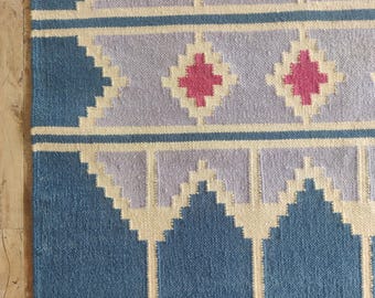 May include: A woven rug with a geometric pattern. The rug features a blue border and sections of blue, lavender, and cream colors. The design includes diamond and triangle shapes in shades of pink and cream.