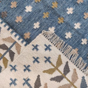 May include: A woven rug featuring a blue field with a repeating pattern of small white and mustard yellow cross shapes. The rug is folded over to reveal a cream-coloured underside with a botanical pattern of blue, olive, and tan leaves and fringe.