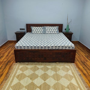 Pu&ograve; includere: Un letto con una struttura in legno, un copriletto con un motivo bianco e blu e due cuscini. Il letto si trova in una stanza con pavimento in legno e una parete di colore blu chiaro. Un tappeto in juta marrone con un motivo a rombi si trova davanti al letto.