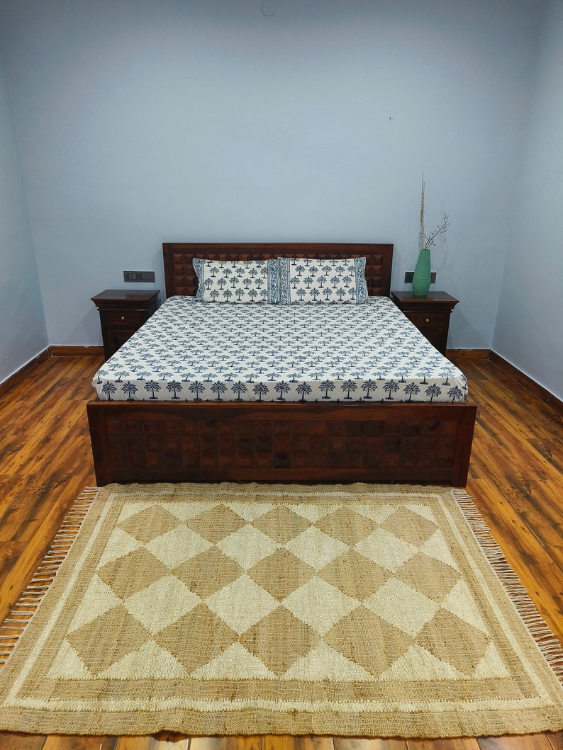 Pu&ograve; includere: Un letto con una struttura in legno, un copriletto con un motivo bianco e blu e due cuscini. Il letto si trova in una stanza con pavimento in legno e una parete di colore blu chiaro. Un tappeto in juta marrone con un motivo a rombi si trova davanti al letto.