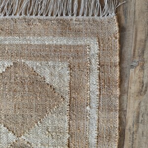 Pu&ograve; includere: Un tappeto in fibra naturale con un motivo geometrico beige e bianco. Il tappeto ha un bordo a frangia.